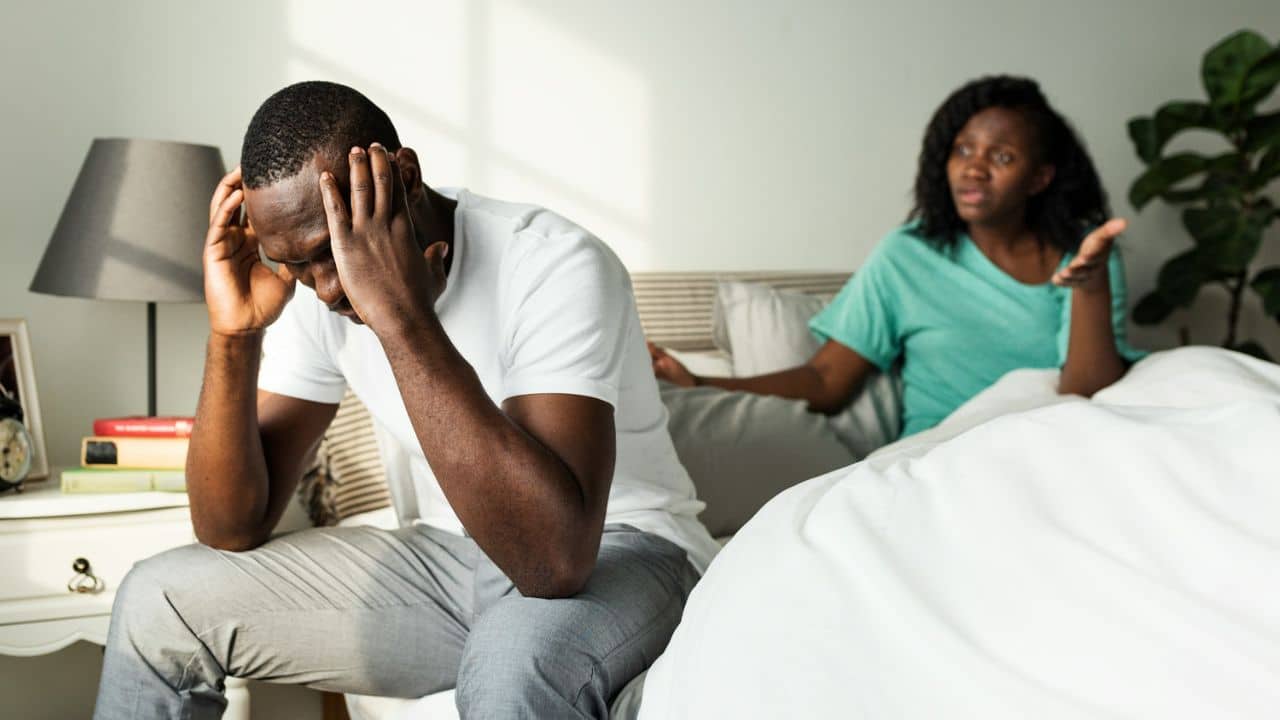 A distressed man sits on a bed holding his head while a woman argues in the background.