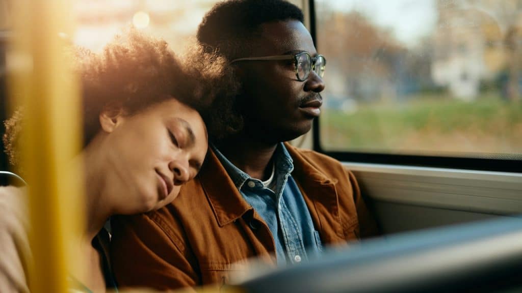 A pensive Black man looking through the window while his girlfriend is falling asleep on his shoulders.