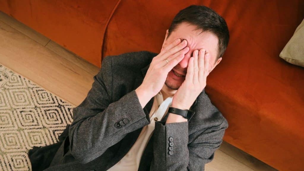 A man in a gray jacket is sitting on the floor, covering his face with his hands.