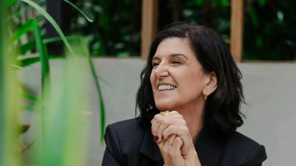 A smiling woman with short dark hair sits in a green, plant-filled setting.