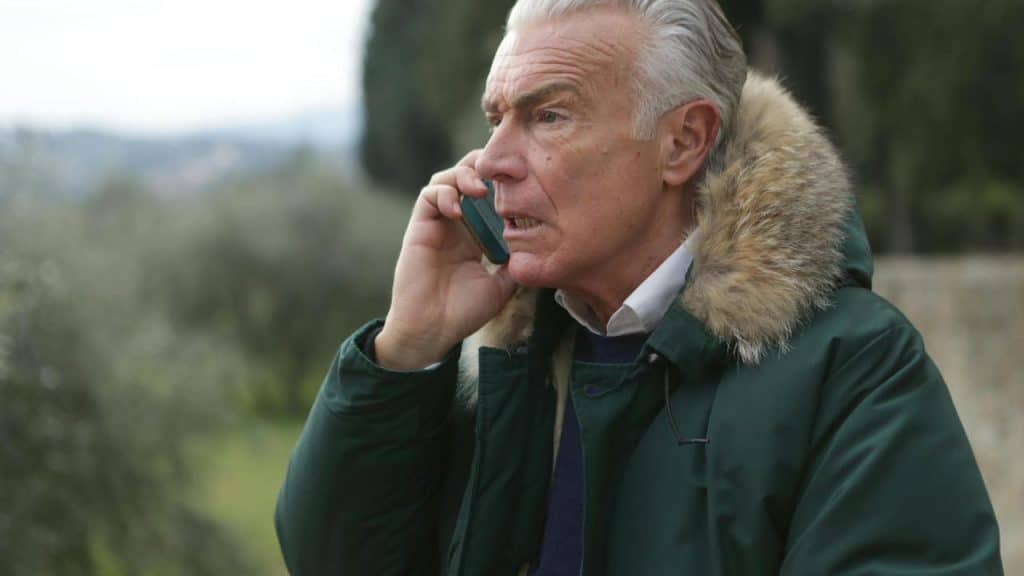 An elderly man wearing a green coat with a fur hood talking on a mobile phone outdoors.