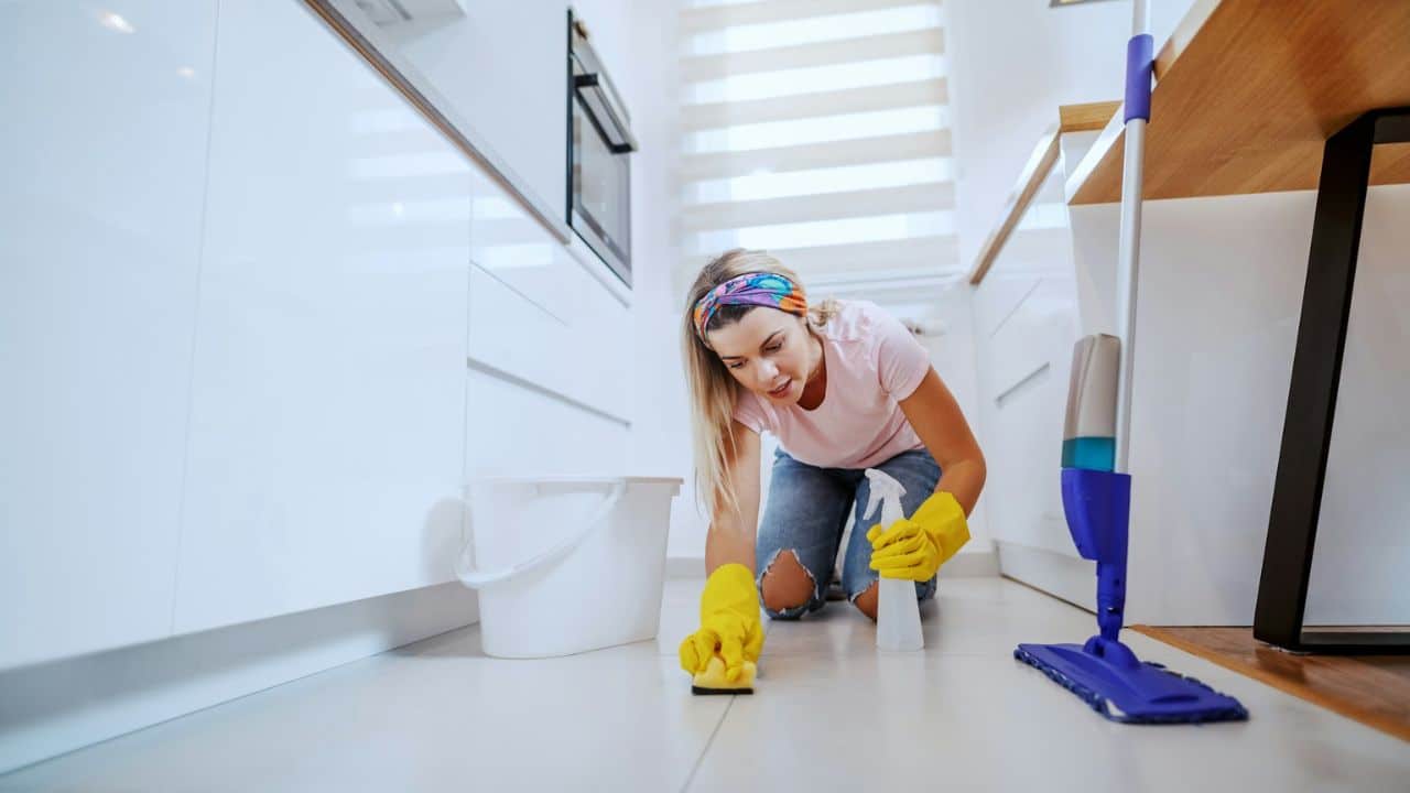 A woman wearing yellow gloves and a colorful headband scrubs a white kitchen floor.