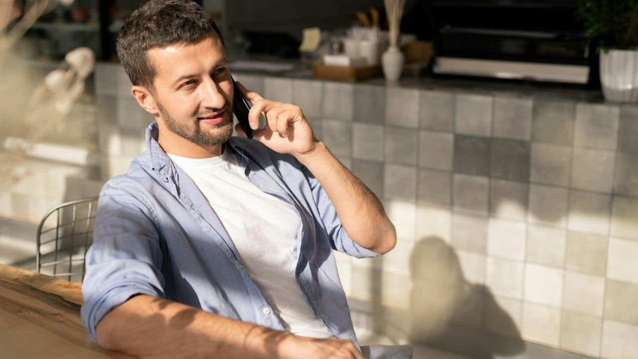 A man with a beard sits at a table talking on a cell phone.