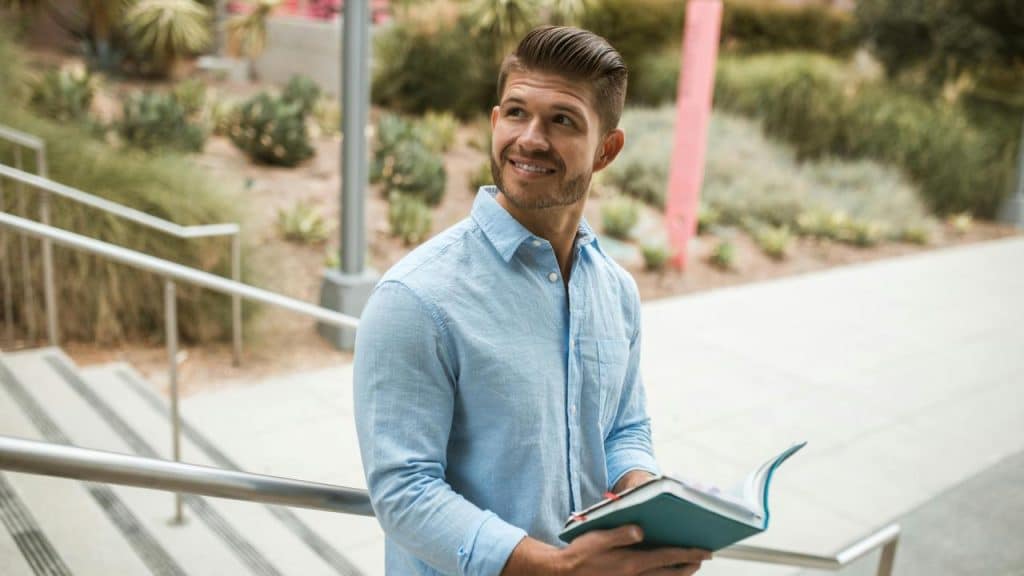 A man in a light blue shirt holding an open book outdoors.