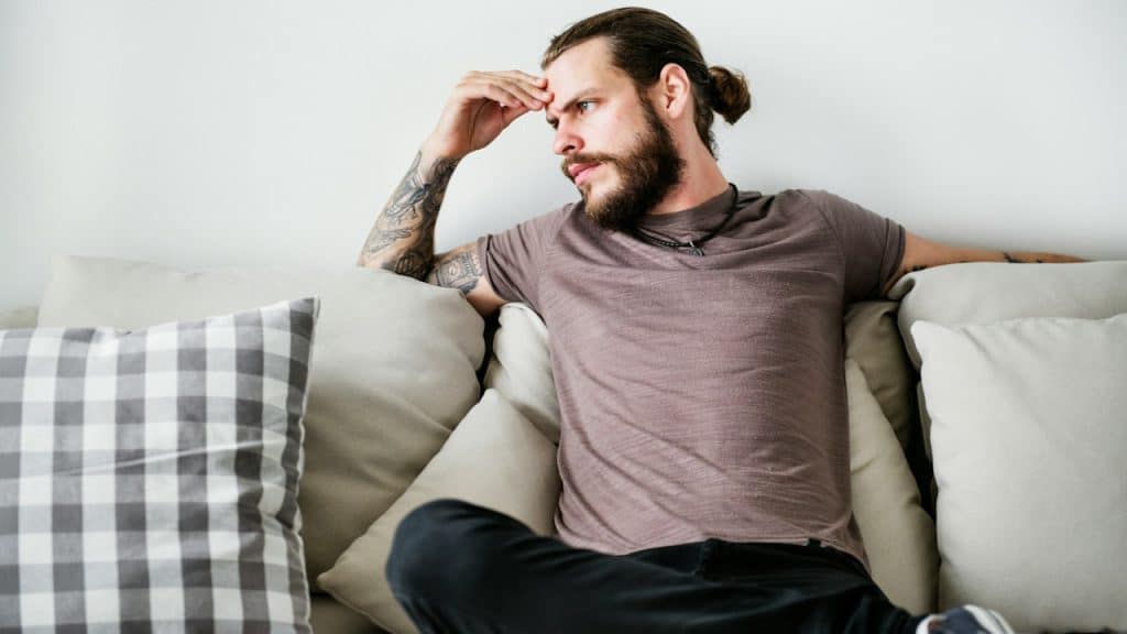 A bearded man with a ponytail sitting on a couch and looking upset.