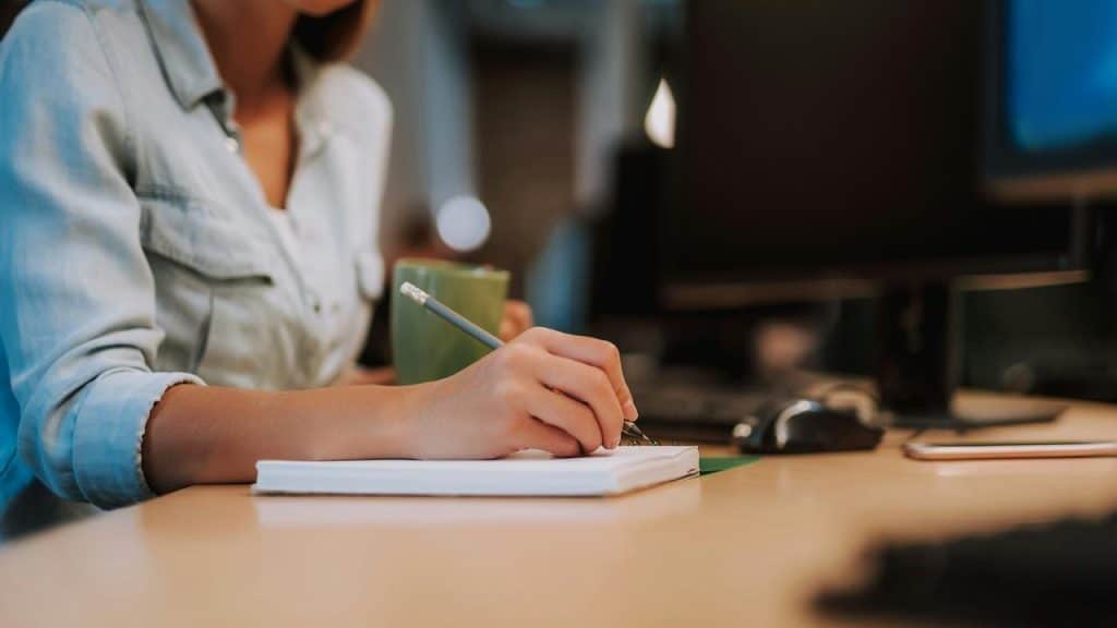 A woman writing in a notebook while holding a mug at her desk.