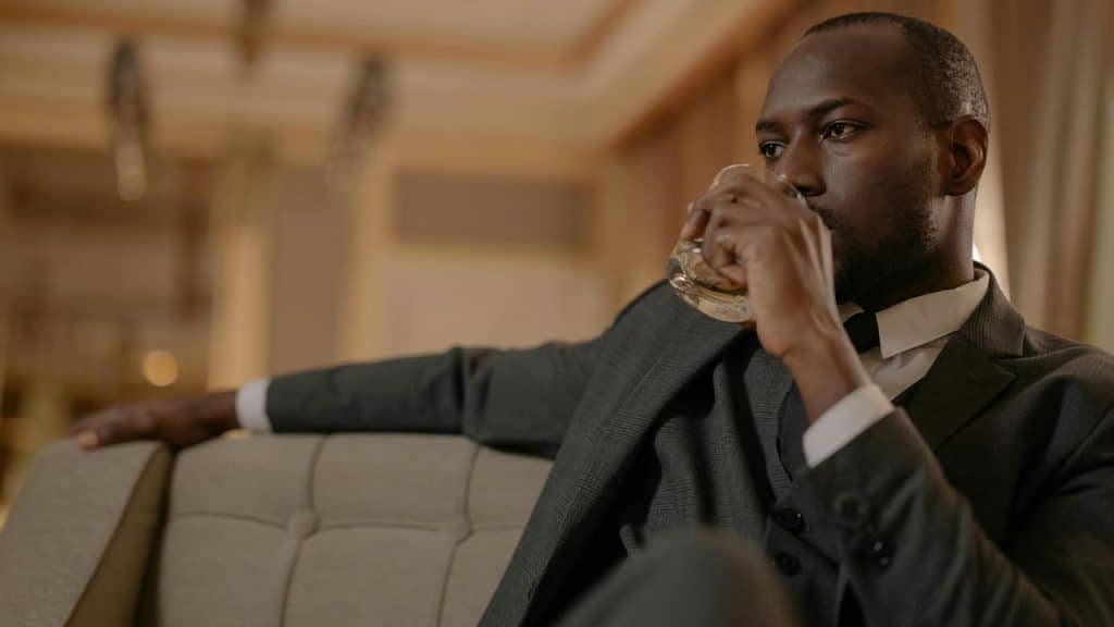 A man in a suit sitting on a couch drinking from a glass.