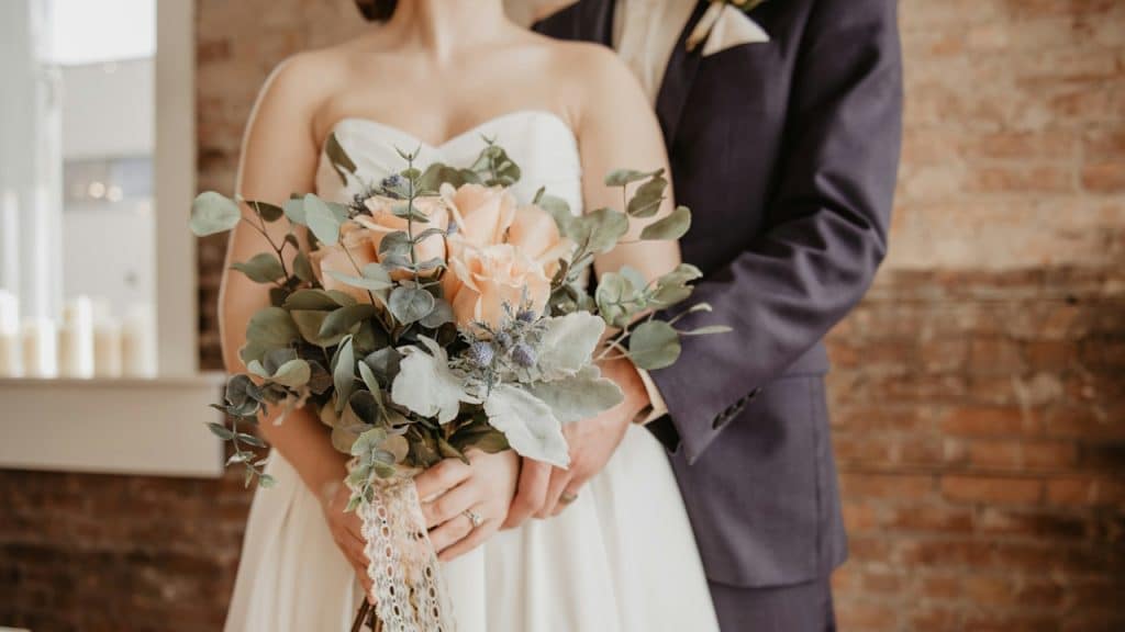 A bride holding a bouquet of peach roses and eucalyptus while standing next to a groom in a navy suit.