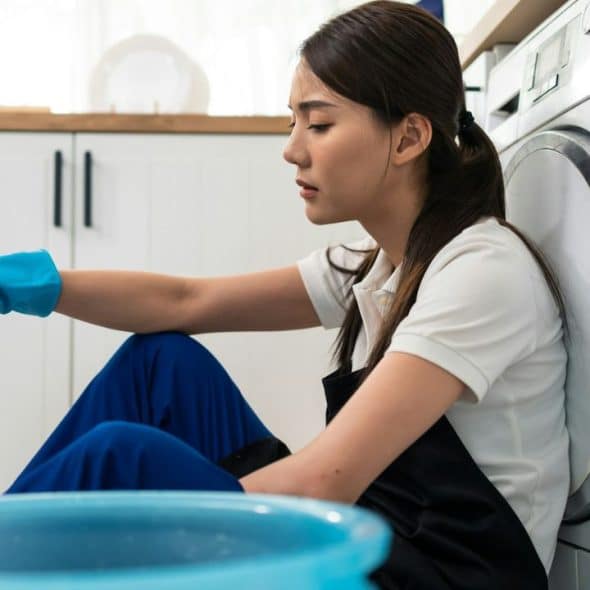 A tired woman with a blue cleaning glove sits on the floor next to a washing machine.