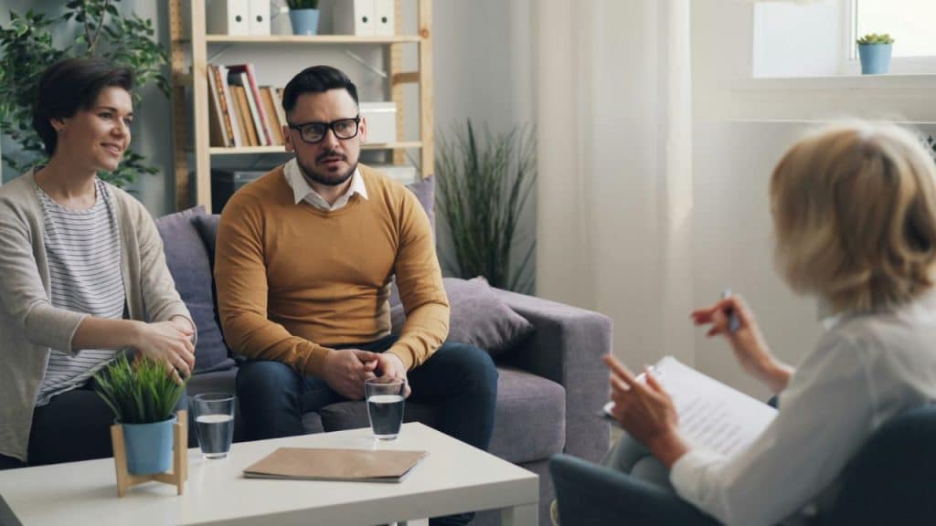 A couple sits on a couch facing a blonde woman taking notes during a consultation.