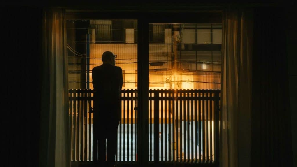 A man stands silhouetted on a balcony at night, looking out at a brightly lit building.