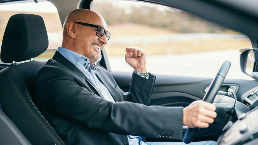 Bald man in a suit and glasses smiling and raising a fist while driving.