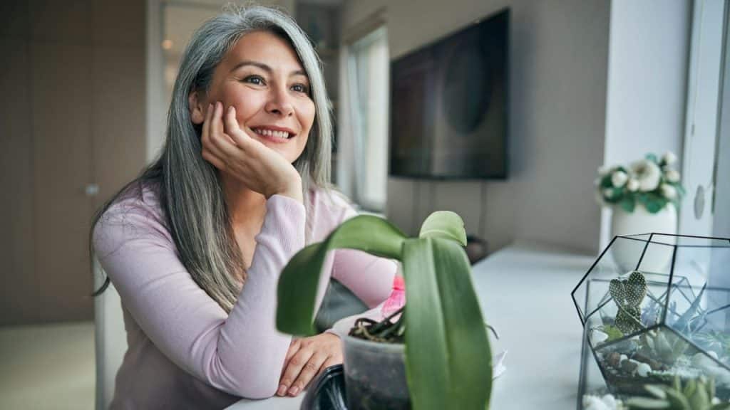Smiling woman with long gray hair resting her chin on her hand indoors.