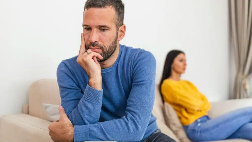 A man in blue sits, looking thoughtful, with a woman in yellow sitting away from him.