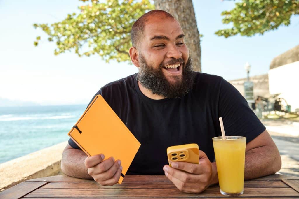 A bearded man holding a notebook in one hand and his phone in the other while sitting at a table by the beach and smiling widely