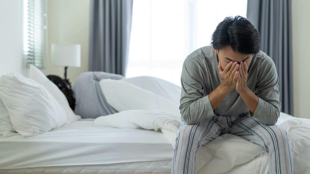Man sitting on bed, face in hands, with a sleeping person behind him.