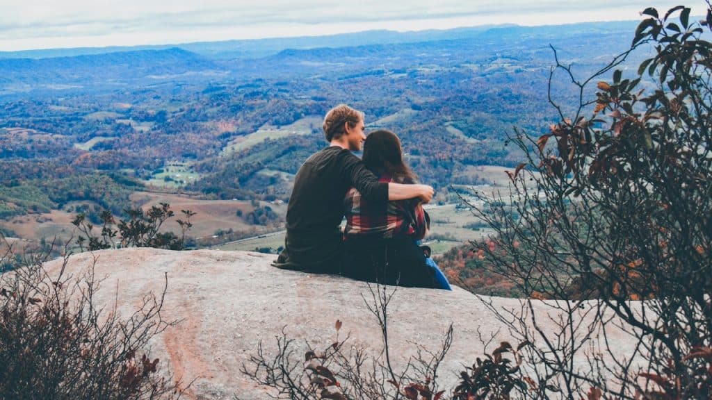 A couple sits on a rocky outcrop with the man's arm around the woman, looking out at a wide, mountainous, green valley view.