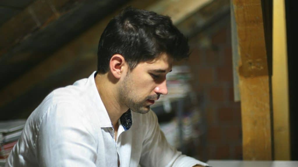 A man in a white shirt is sitting in a dimly lit attic, looking down.