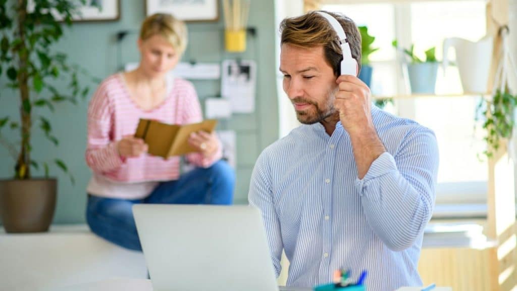 Man wearing headphones uses a laptop while a woman reads in the background.