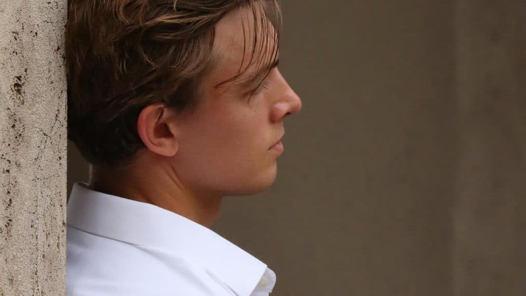 A young man in a white shirt leaning against a wall, looking thoughtful.