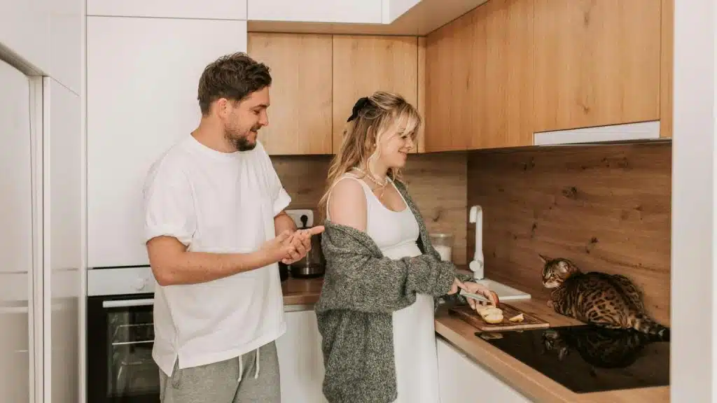A couple preparing food in the kitchen while a cat sits on the counter watching.