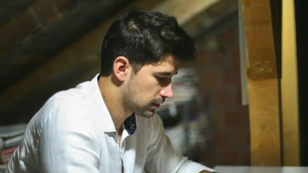 A man in a white shirt is sitting in a dimly lit attic, looking down.
