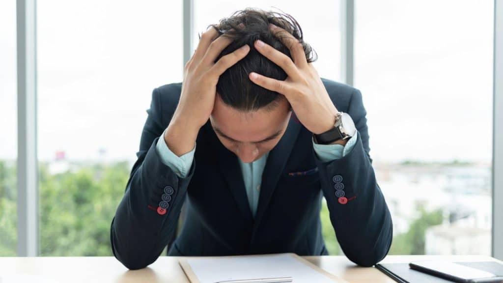 A man in a suit is sitting at a desk, holding his head with his hands in frustration.