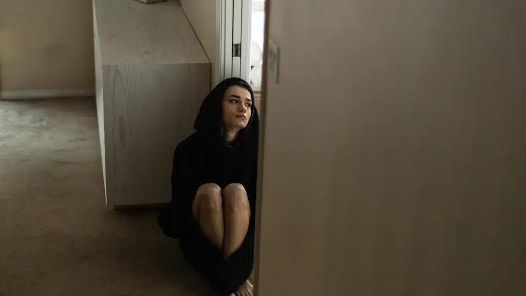A woman in a dark hoodie sits on the floor looking thoughtful.
