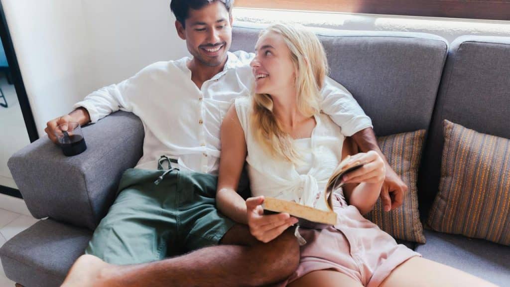 A smiling couple relaxes on a couch looking at each other while smiling.