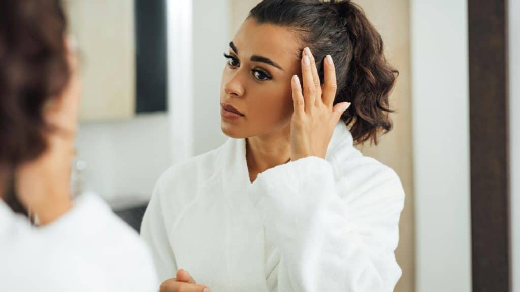 A woman in a white bathrobe looking at herself in the mirror and touching her hair.
