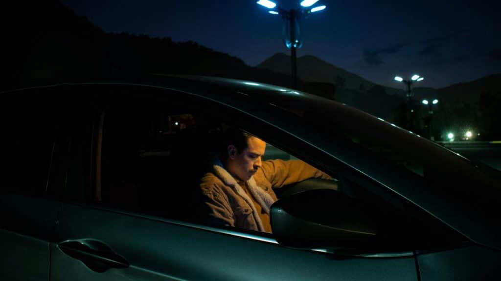 A man sitting in a parked car at night, illuminated by dim streetlights.