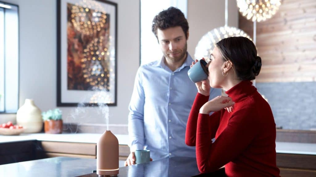 A woman in a red turtleneck drinks from a mug next to a man and a diffuser.