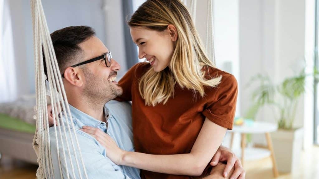 Smiling man in glasses and woman in an orange top embracing while sitting in a white indoor swing chair.