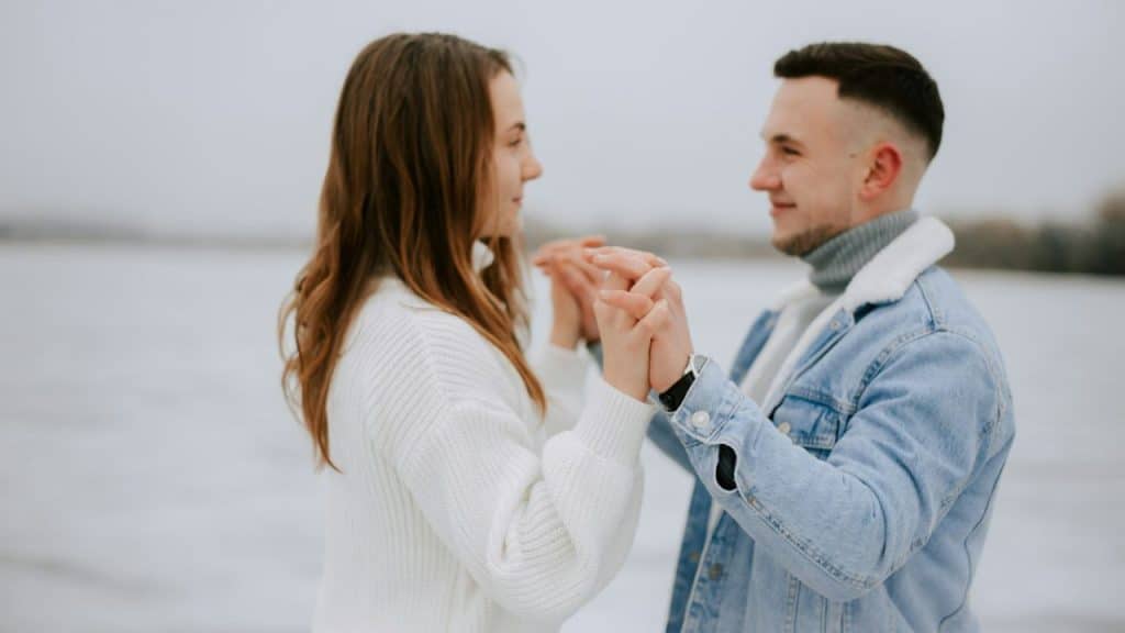 Man and woman holding hands and facing each other outdoors in a cold, white setting.