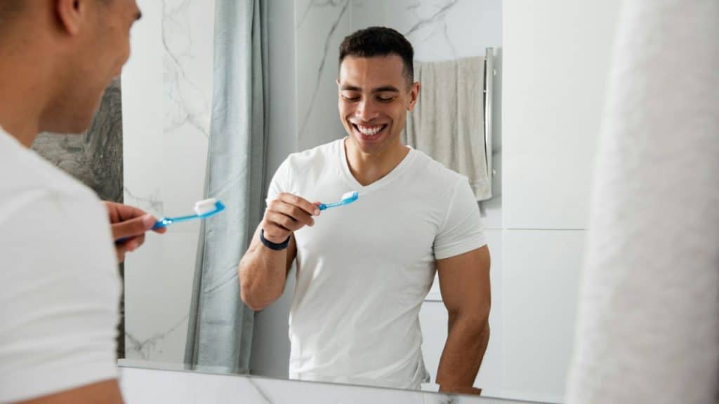 Smiling man in a white shirt holding a toothbrush, reflected in a bathroom mirror.