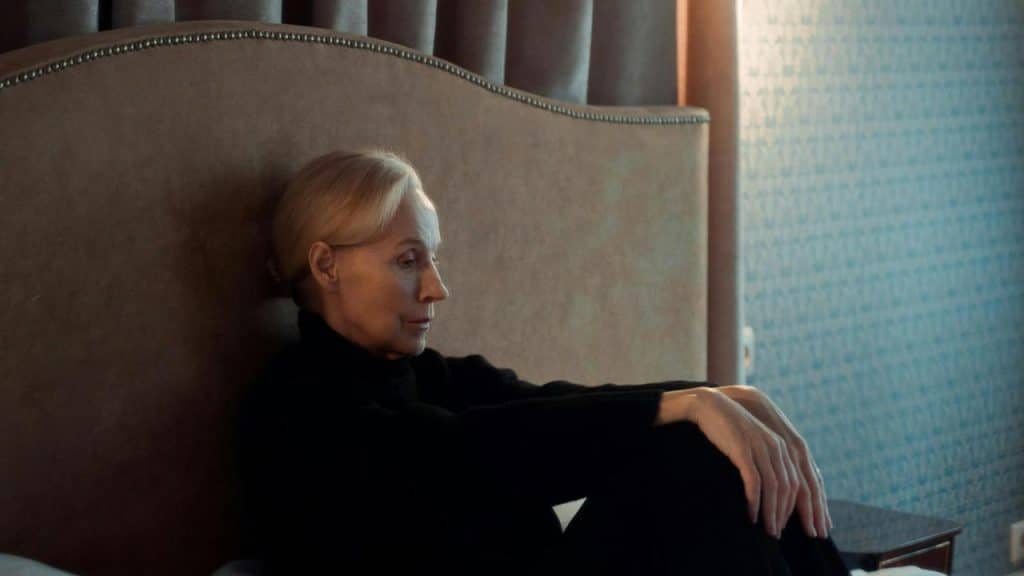 An elderly woman sitting on a bed looking thoughtful.