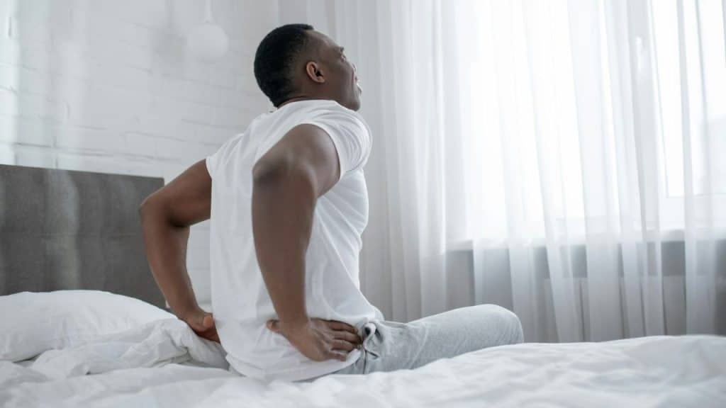 A man sits on a bed, holding his lower back with both hands due to pain.