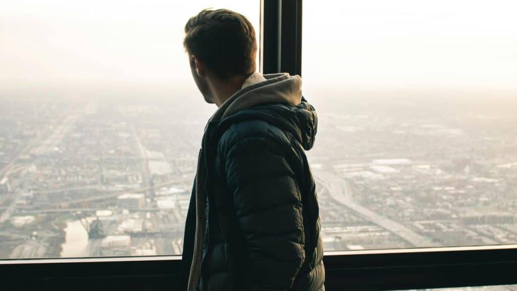 A man in a jacket looks out a tall window at a cityscape below.