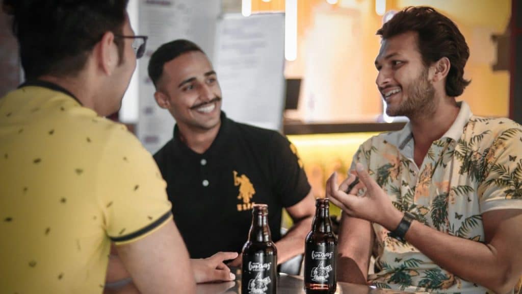 Three men smiling, sitting at a table with two dark bottles, talking indoors.