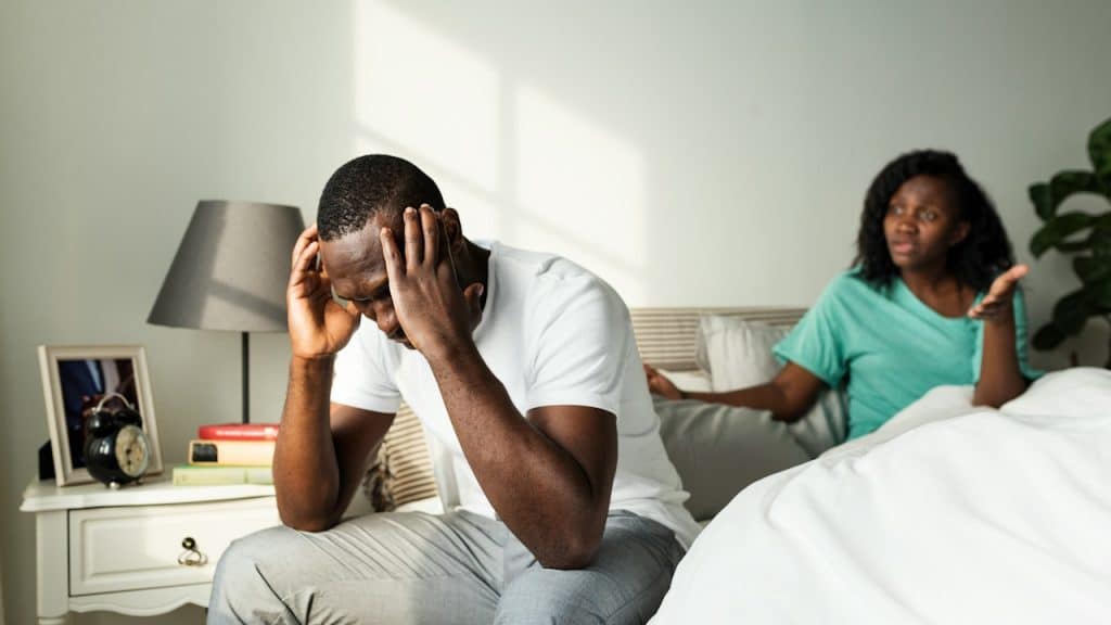 A man holding his head in his hands and looking stressed while sitting on a bed with a woman in the background who’s telling him something.