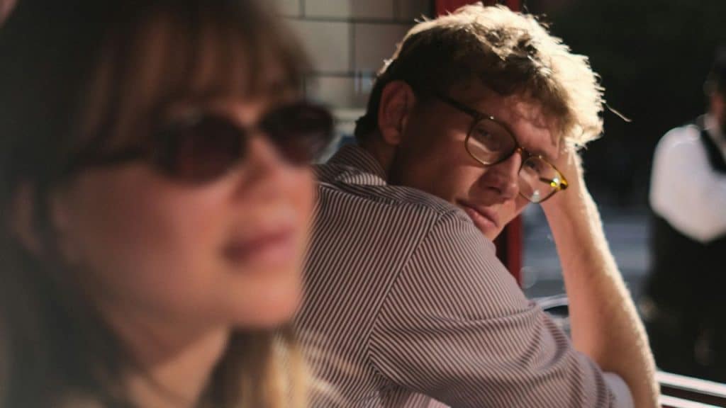 A man wearing glasses looks over his shoulder past a blurry woman in sunglasses.