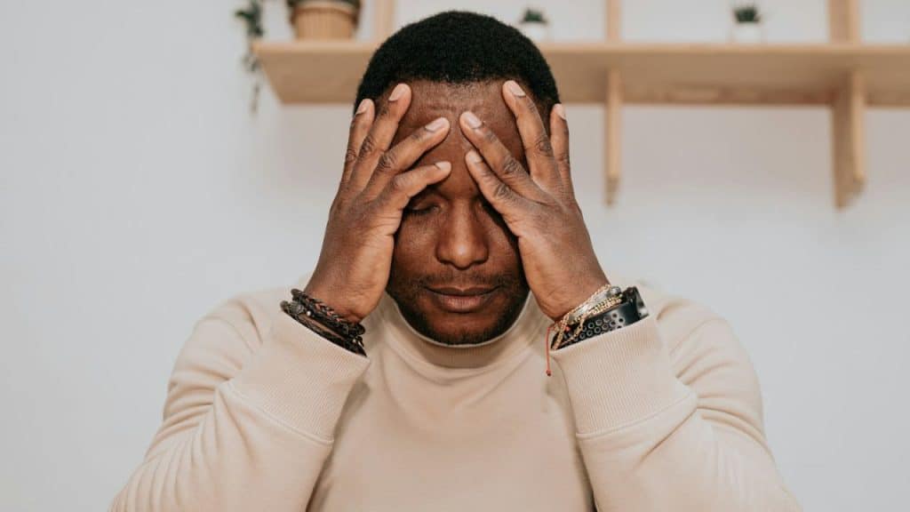 A distressed man in a tan sweatshirt holds his hands to his temples.
