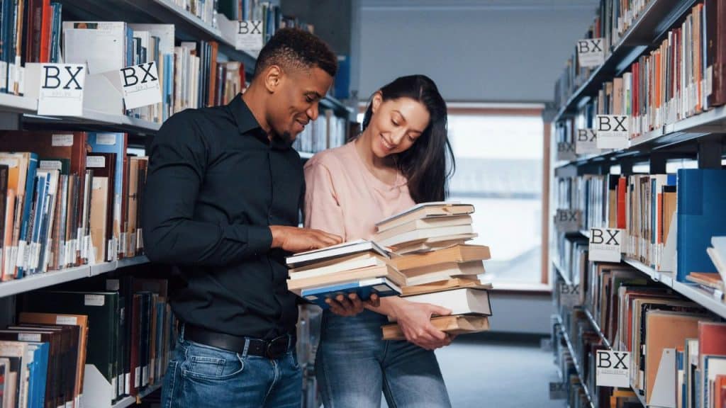 A smiling young man and woman are standing in a library aisle, holding and looking through stacks of books.