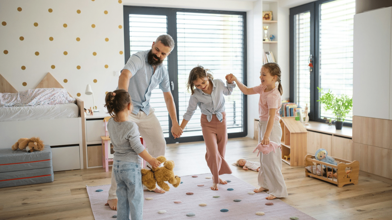 A father and his three daughters playing together on a rug in a sunlit room.