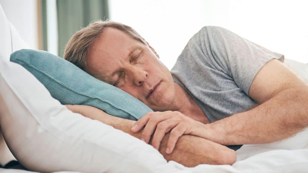 A middle-aged man in a grey shirt is sleeping peacefully on a blue pillow.