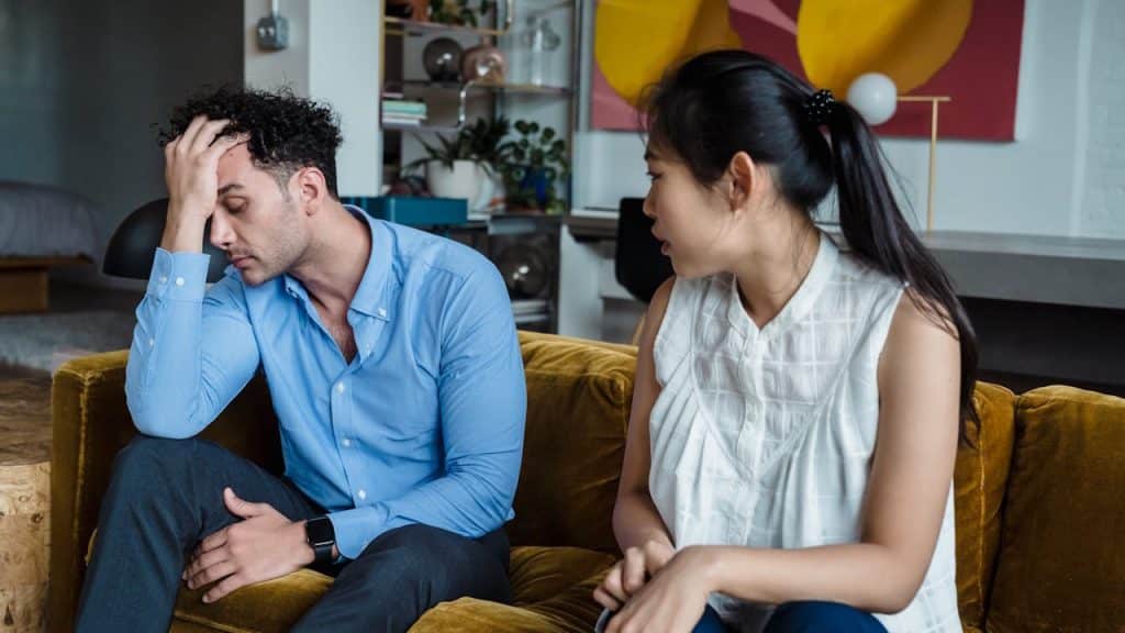 A couple sitting on a couch, with the man looking distressed and holding his forehead.