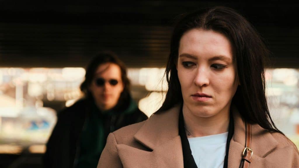 Close-up of a somber woman in a tan coat with a blurred person in the background.