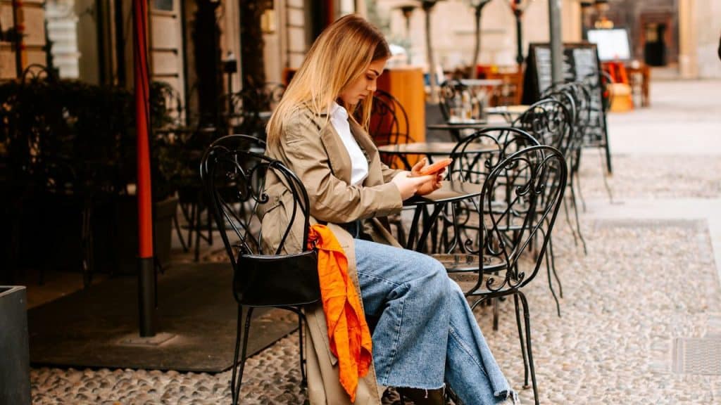 Woman in a tan jacket and jeans sitting at an outdoor cafe table, looking at her phone.