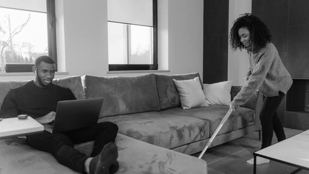 A black and white image of a man sitting on a sofa working on a laptop while a woman vacuums the floor beside him.