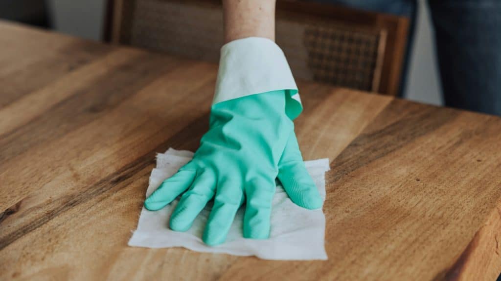 Close-up of a hand in a green and white cleaning glove wiping a wooden table with a white cloth.